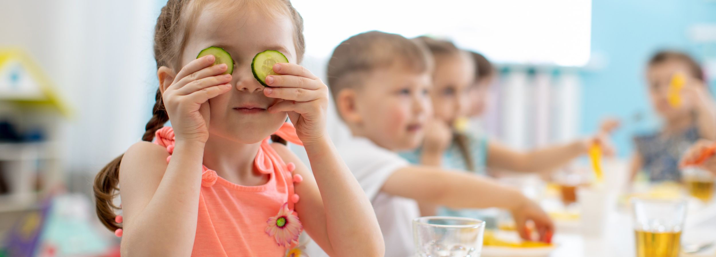 Hero-Image Menü Niederwiesa GmbH & Co. KG Kinder am Esstisch, im Vordergrund Mädchen mit Gurkenscheiben